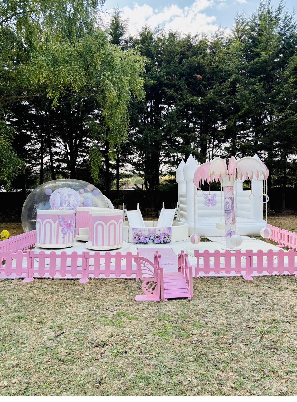 Pink and white butterfly-themed setup with teacup carousel, Bubble House Dome, ball pit with slides, carousel tree and white bouncy castle, with a pink bridge entrance